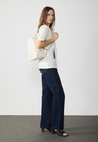 Beige steppelt hátizsák, amelynek formája rendezett, állítható pántokkal és aranyszínű részletekkel rendelkezik. A modellen fehér grafikus póló és sötét farmernadrág van.