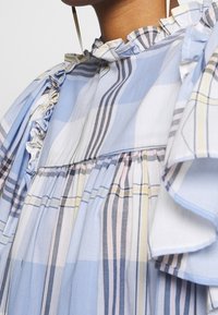 Detailní záběr na zmačkané pastelově kostkované šaty s modrými, žlutými a růžovými pruhy na horní části hrudníku a rameně osoby.