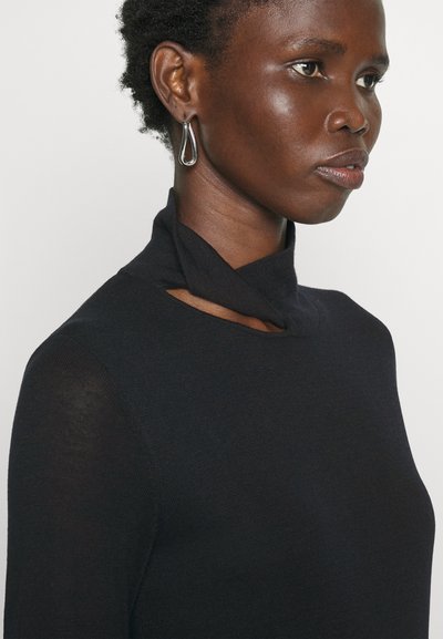 Μαύρο εφαρμοστό μπλουζάκι με σχέδιο μισού λαιμού, που διαθέτει λεπτομέρεια με ανοίγματα στον γιακά. Λείο υλικό. Το μοντέλο φορά ασημένια σκουλαρίκια σε σχήμα δάκρυ.