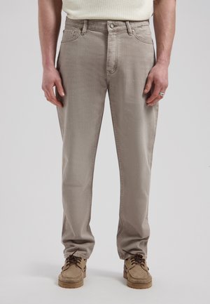 Man draagt beige slim-fit jeans, wit gestructureerd overhemd, bruine suède schoenen en een zilveren ring aan zijn rechterhand, staand tegen een effen achtergrond.