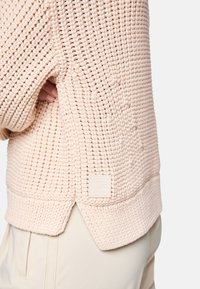 Light pink knitted sweater featuring a textured pattern, ribbed cuffs, and a rectangular logo patch at the lower left side.