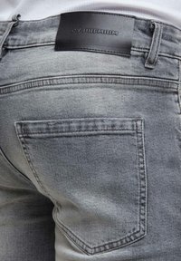 Jean en denim gris avec des coutures visibles, une poche arrière, des passants pour ceinture et une étiquette de marque en cuir noir indiquant "GY PREMIUM."