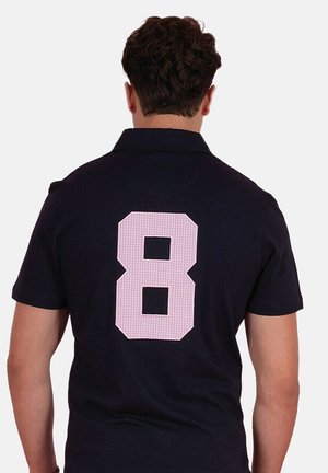 Homme aux cheveux bouclés foncés portant une chemise noire à manches courtes avec un grand chiffre 8 à carreaux roses dans le dos.