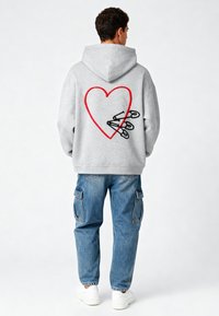 Felpa grigia con cappuccio caratterizzata da un design di cuore rosso con spille di sicurezza nere sul retro, abbinata a jeans cargo blu e sneakers bianche.