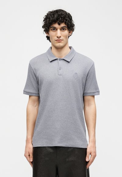 Νεαρός με σγουρά μαλλιά που φοράει ένα ανοιχτό γκρι πόλο με κοντά μανίκια και μαύρο υφασμάτινο παντελόνι, στέκεται μπροστά σε ένα απλό λευκό φόντο.