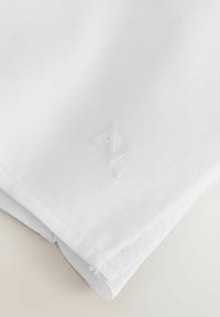 Tissu blanc plié avec un logo "Z" subtilement embossé près d'une bordure cousue sur une surface claire.