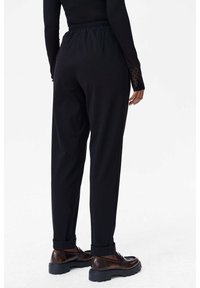 Zwarte taps toelopende broek met een elastische tailleband en opgerolde manchetten, gecombineerd met bruine schoenen. De stof is glad en heeft een gestructureerde pasvorm.