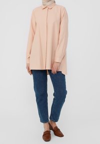 Camicia a maniche lunghe di un delicato color pesca con colletto e chiusura a bottoni, abbinata a jeans blu medio e mocassini marroni. Tessuto di trama liscia.