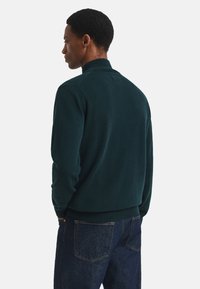Dunkelgrüner Strickpullover mit hohem Kragen. Das Design verfügt über lange Ärmel und gerippte Bündchen, kombiniert mit blauen Jeans.
