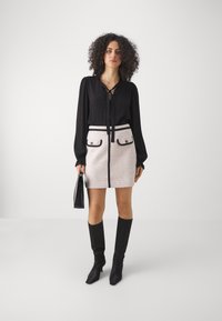 Blouse noire à manches longues avec détail de nœud, associée à une mini-jupe en tweed rose et gris avec des accents noirs et deux poches avant. Des bottes noires jusqu'aux genoux complètent le look.