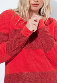 Czerwony sweter dziewiarski z długim rękawem w paski, z różnymi odcieniami czerwieni i teksturowanym wzorem. Klasyczny ściągacz przy dekolcie i mankietach.