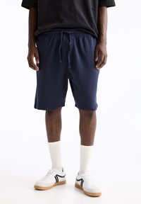 Shorts de sudation bleu marine avec une taille élastique et un cordon de serrage. Associés à des chaussettes blanches et des baskets avec des accents noirs.