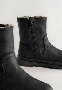 Svarta ankelskor med sidodragkedjor och pälsfoder, som visar strukturerat material och tjocka gummisålar på en enfärgad bakgrund.