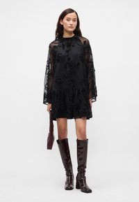 Crna haljina s cvjetnim vezenjem, prozirnim dugim rukavima i volanima na rubu. Uparena s čizmama do koljena od sjajne burgundske boje i odgovarajućom torbicom.