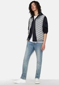 Lätt grå puffer väst med quiltat mönster, lager på lager över en svart långärmad tröja och en vit t-shirt, matchat med blå jeans och vita sneakers.