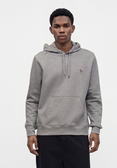 Jeune homme portant un sweat à capuche gris avec un petit écusson zèbre sur la poitrine, debout devant un fond blanc uni.
