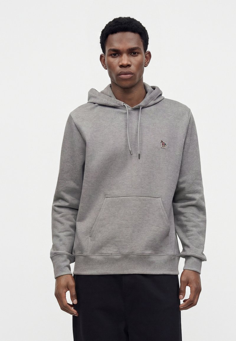 Jeune homme portant un sweat à capuche gris avec un petit écusson zèbre sur la poitrine, debout devant un fond blanc uni.