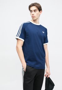 Námornícky modré tričko s bielymi akcentmi a tromi bielymi pruhmi na rukávoch; má malú logo na hrudi. Textúrovaná, džersejová tkanina.