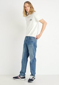 Hvid kortærmet t-shirt med "Lee" logo, parret med blå denim jeans og sorte sneakers med hvide og røde accenter.