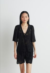 Blusa nera a occhietti con maniche corte e scollatura a V, caratterizzata da un orlo scallop e un motivo simile a pizzo, abbinata a pantaloni corti coordinati.