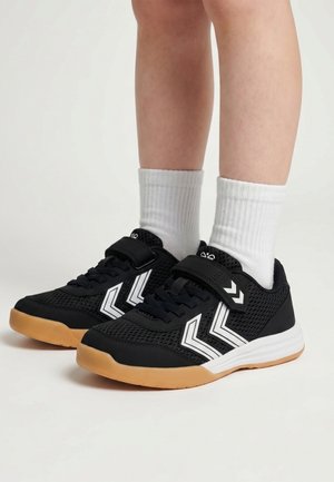 Kind trägt schwarz-weiße Sportschuhe mit Gummisohlen und weißen Socken und steht vor hellem Hintergrund.