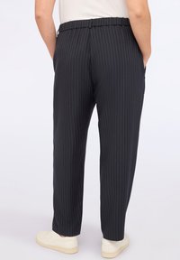 Osoba w czarnych spodniakach w prążki i białych sneakersach, stojąca z rękami w kieszeniach, widok z tyłu na jednolity jasnym tle.