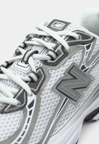 Zapatilla New Balance blanca y gris con parte superior de malla, cordones blancos y logo de la marca en el costado y en la lengüeta, mostrada en primer plano.