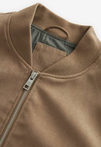 Veste bomber en daim marron avec col côtelé et fermeture éclair argentée partiellement fermée, laissant apparaître la doublure intérieure grise et une boucle pour suspendre.