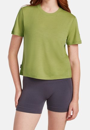 Person iført en kortærmet grøn T-shirt og højtaljede mørkegrå tætsiddende shorts mod en ensfarvet baggrund.