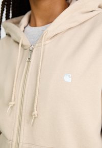 Beige zip-up hette med snøring på hetten. Har en liten, skinnende logo på venstre bryst. Mykt bomullsmateriale med en jevn tekstur.