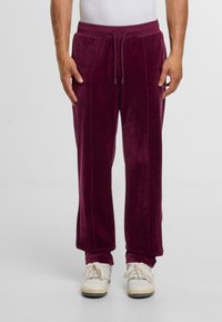 Mann in bordeauxroten Velourhosen mit Vordernähten und weißen Turnschuhen, der vor einem schlichten hellen Hintergrund steht.