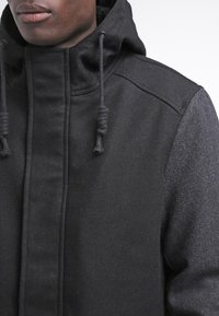 Schwarze Kapuzenjacke aus einer Mischung aus glattem und strukturiertem Material, mit verstellbaren Kordeln und hohem Kragen für zusätzlichen Wärme.