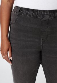 Zwarte denim broek met een elastische tailleband, voorzien van zijzakken en een gladde textuur. Close-up van de taille en hand, met nadruk op de pasvorm.