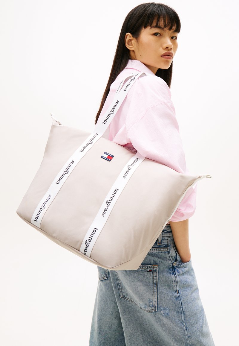 Τσ άντα ώμου σε ανοιχτό μπεζ χρώμα με μεγάλο, ορθογώνιο σχήμα. Διαθέτει εμφανείς λευκές λουράκια με το κείμενο "tommyjeans" και ένα μικρό λογότυπο.