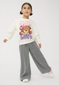 Kremowa bluza z grafiką niedźwiedzia i napisem; szare szerokie spodnie; miękka tkanina; prezentowana w stojącej pozycji na jasnym tle.