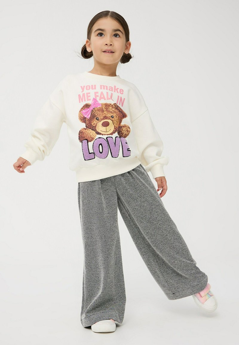 Kremowa bluza z grafiką niedźwiedzia i napisem; szare szerokie spodnie; miękka tkanina; prezentowana w stojącej pozycji na jasnym tle.