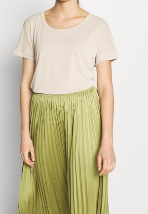 Vrouw die een beige korte mouw top draagt, ingestopt in een lichtgroene geplooide rok met hoge taille, staand tegen een effen achtergrond.