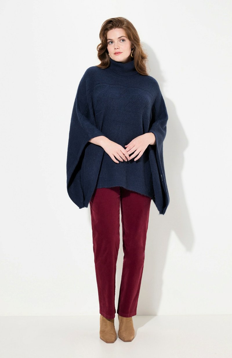Poncho en tricot bleu marine avec un col haut et un motif texturé, associé à un pantalon bordeaux et des bottines beige.