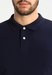 Polo en bleu marine en tissu texturé avec un col classique, comportant deux boutons blancs et des détails de logo subtils.
