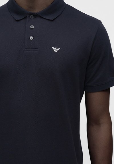 Polo bleu marine foncé avec trois boutons et un petit logo blanc sur la poitrine gauche, porté par une personne à la peau foncée.