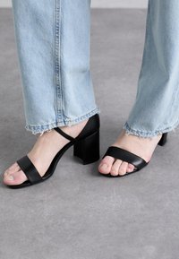 Sandali neri con tacco a spillo, punta quadrata e due cinturini. Tacco alto a blocco e design aperto, abbinati a jeans blu chiaro con orlo sfrangiato.