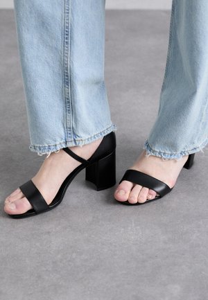 Sandali neri con tacco a spillo, punta quadrata e due cinturini. Tacco alto a blocco e design aperto, abbinati a jeans blu chiaro con orlo sfrangiato.