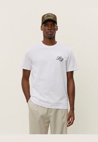 Camiseta de algodón blanca de manga corta y escote redondo, con un pequeño logotipo bordado en negro "LD" en el lado izquierdo del pecho.
