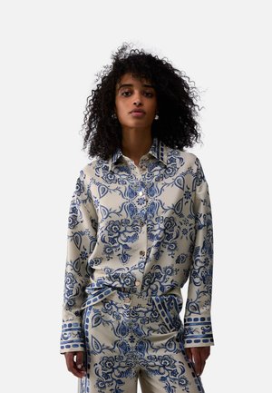 Giovane donna con capelli ricci che indossa una camicia e pantaloni beige con motivi floreali e paisley blu.