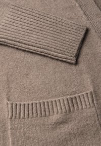 Cardigan en tricot beige avec des manches côtelées et une poche, fabriqué en laine douce, avec une texture lisse et des variations de couleur subtiles.