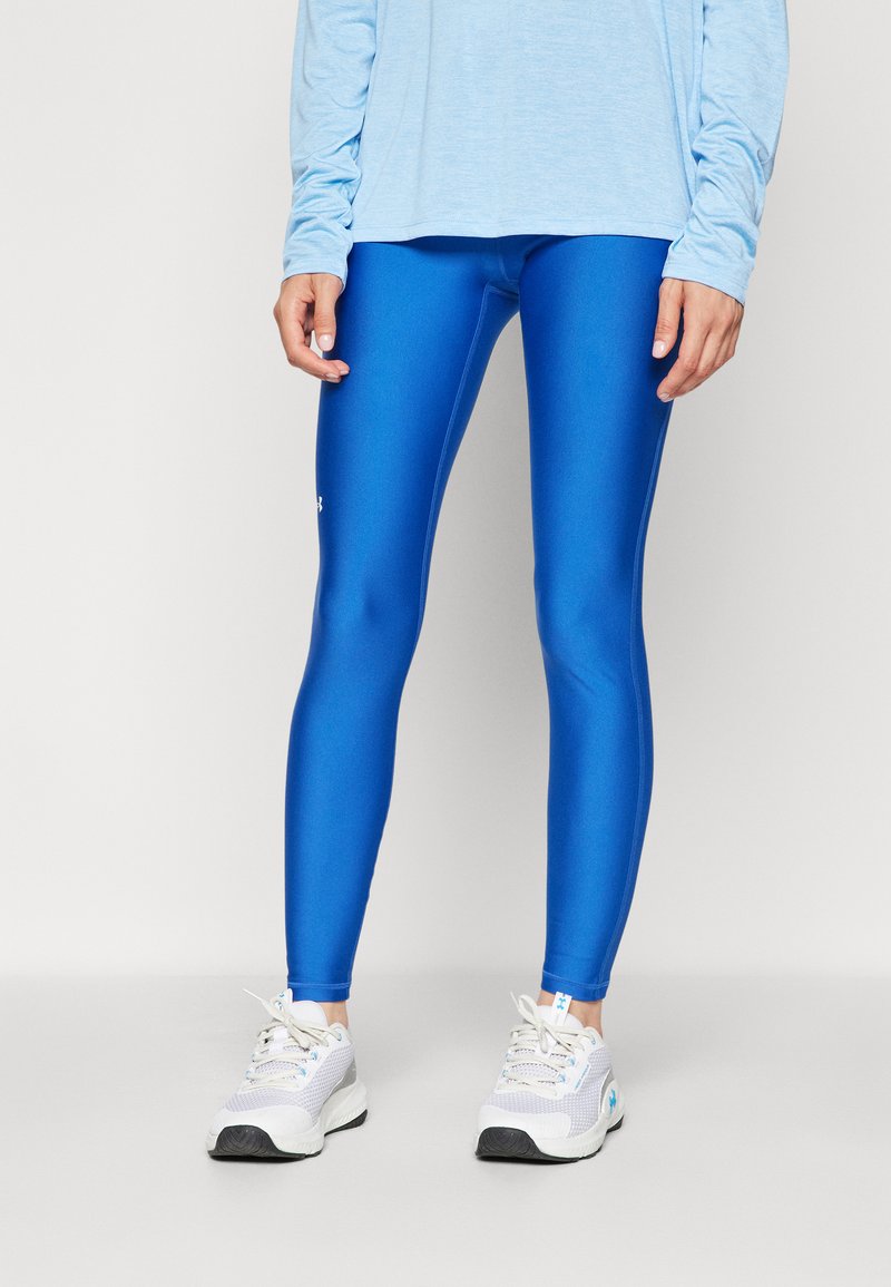 Leggings blu, lucidi, con una texture liscia e un design aderente, abbinati a una maglietta a maniche lunghe di un blu chiaro e scarpe da ginnastica grigie.