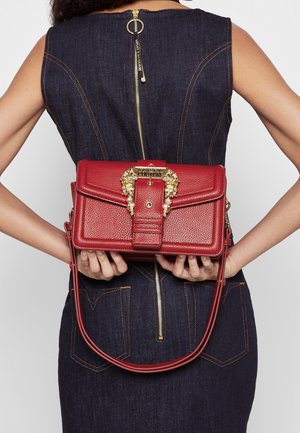 Persoon in een donker denimjurk met een gouden rits houdt achter hun rug een rode leren handtas vast met een gouden decoratieve leeuwenkopsluiting.