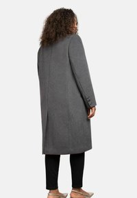 Cappotto in lana grigia con taglio dritto, maniche lunghe e polsini con bottoni. Presenta una fessura centrale sul retro per facilitare i movimenti.