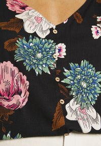 Černá květinová blůza s velkými růžovými a modrými květy, dřevěné knoflíky a dekoltem do V. Tkanina vypadá hladce s mírným leskem.