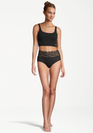 Frau trägt schwarzes kurzes Oberteil und hoch taillierte Unterwäsche mit Spitzenbund, steht barfuß vor weißem Hintergrund.